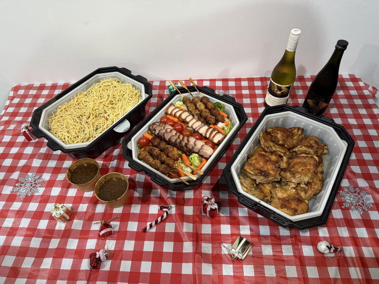 Three self-heating trays of noodles, satay and chicken on a chequered tablecloth